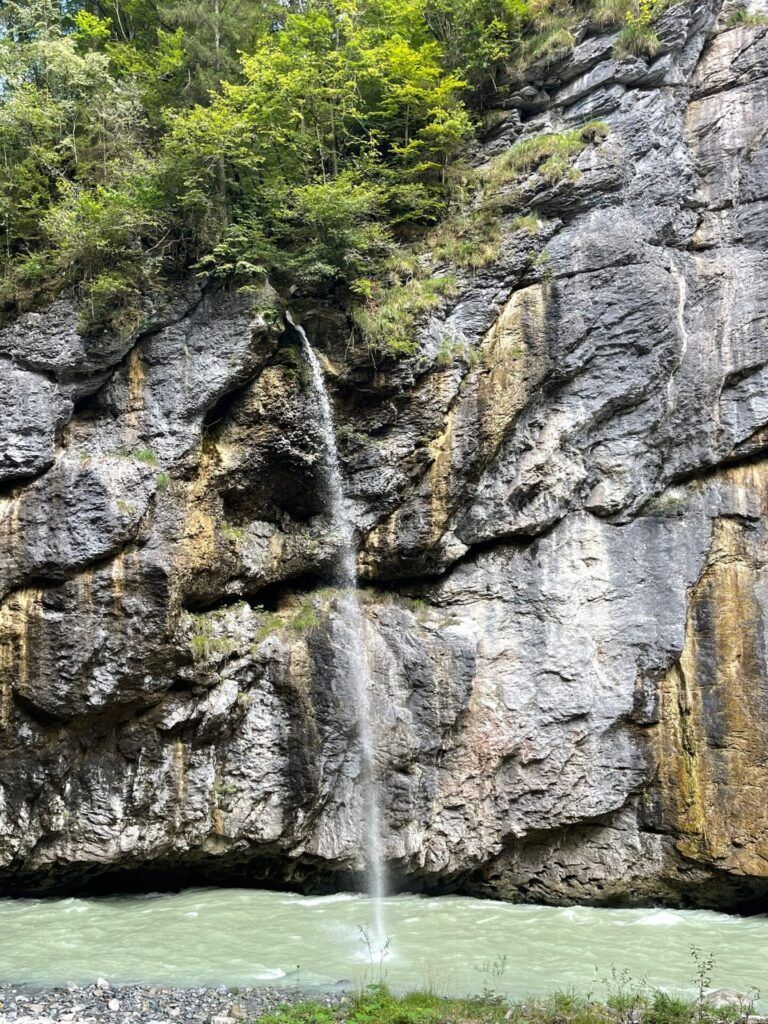 A small waterfall cascading down a rocky cliff surrounded by greenery, with a river flowing at the base.