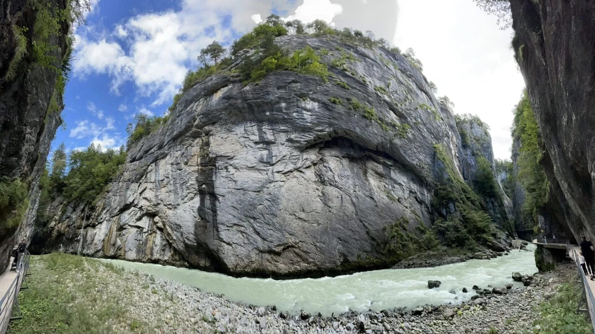 A panoramic view of Aare Gorge, featuring steep rock faces and a flowing river at the bottom, under a blue sky with scattered clouds.