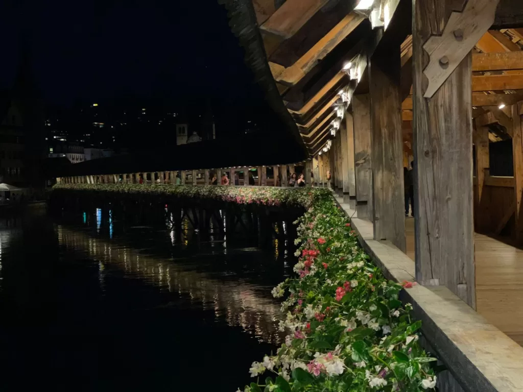 A view of Chapel Bridge in Lucerne at night, illuminated by lights, with flowers along the wooden walkway and reflections in the water.