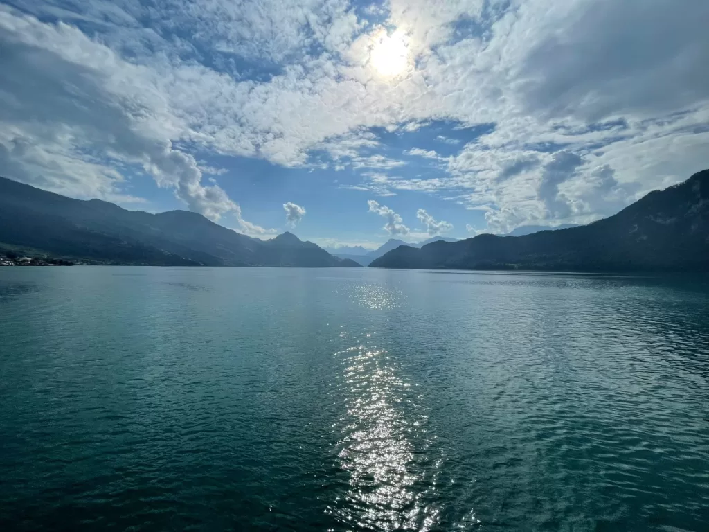 A serene view of a calm lake surrounded by mountains under a partly cloudy sky, with sunlight reflecting off the water's surface.