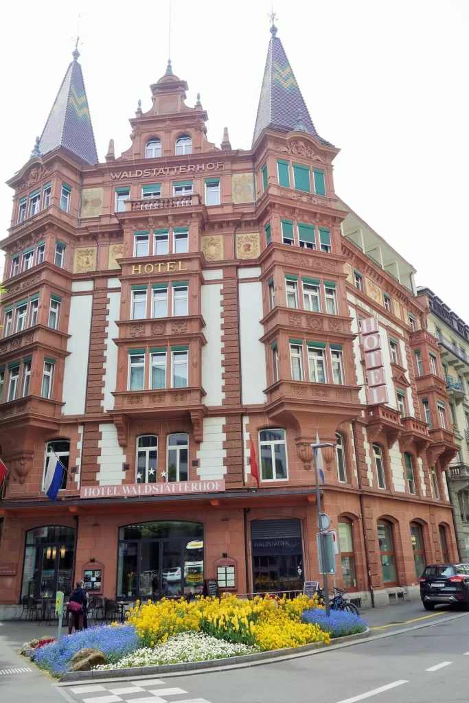 Exterior view of the Hotel Waldstätterhof in Lucerne, Switzerland, showcasing its distinctive red facade, intricate architectural details, and colorful flower beds in front.