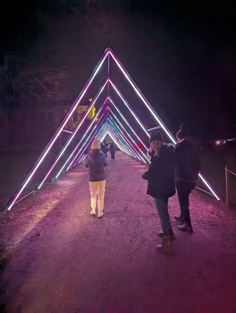 A colorful illuminated light tunnel with triangular shapes along a path at night, with people walking through it.