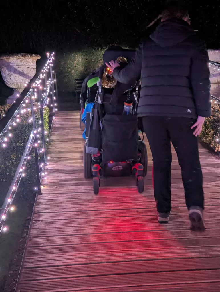 A powerchair from behind driving along a wooden pathway adorned with twinkling Christmas lights, set in a festive outdoor environment.