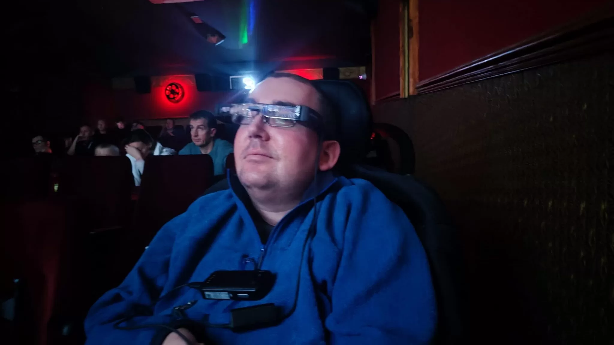 A person seated in a cinema wearing WatchWord subtitle glasses, with a relaxed expression while watching a film.