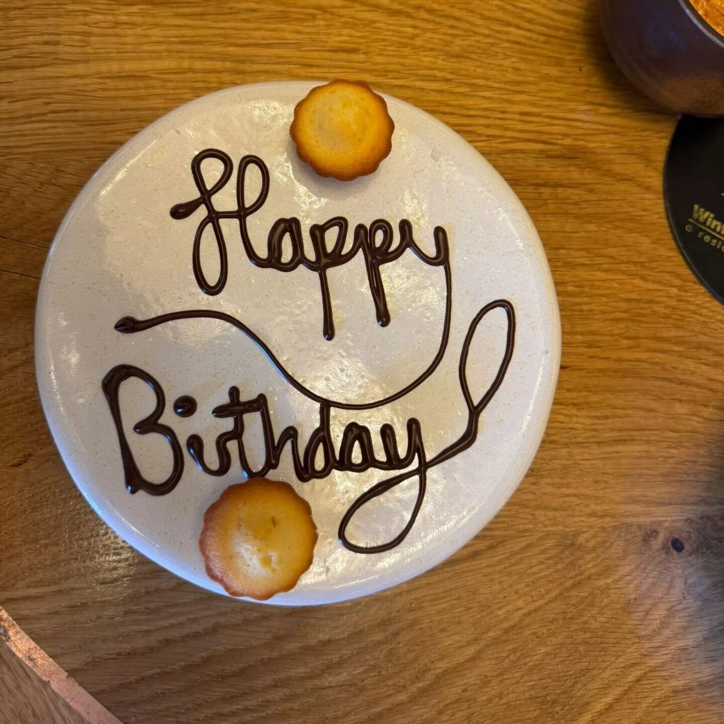A round plate with the words 'Happy Birthday' written in chocolate, accompanied by a small cake on the side, set on a wooden table.