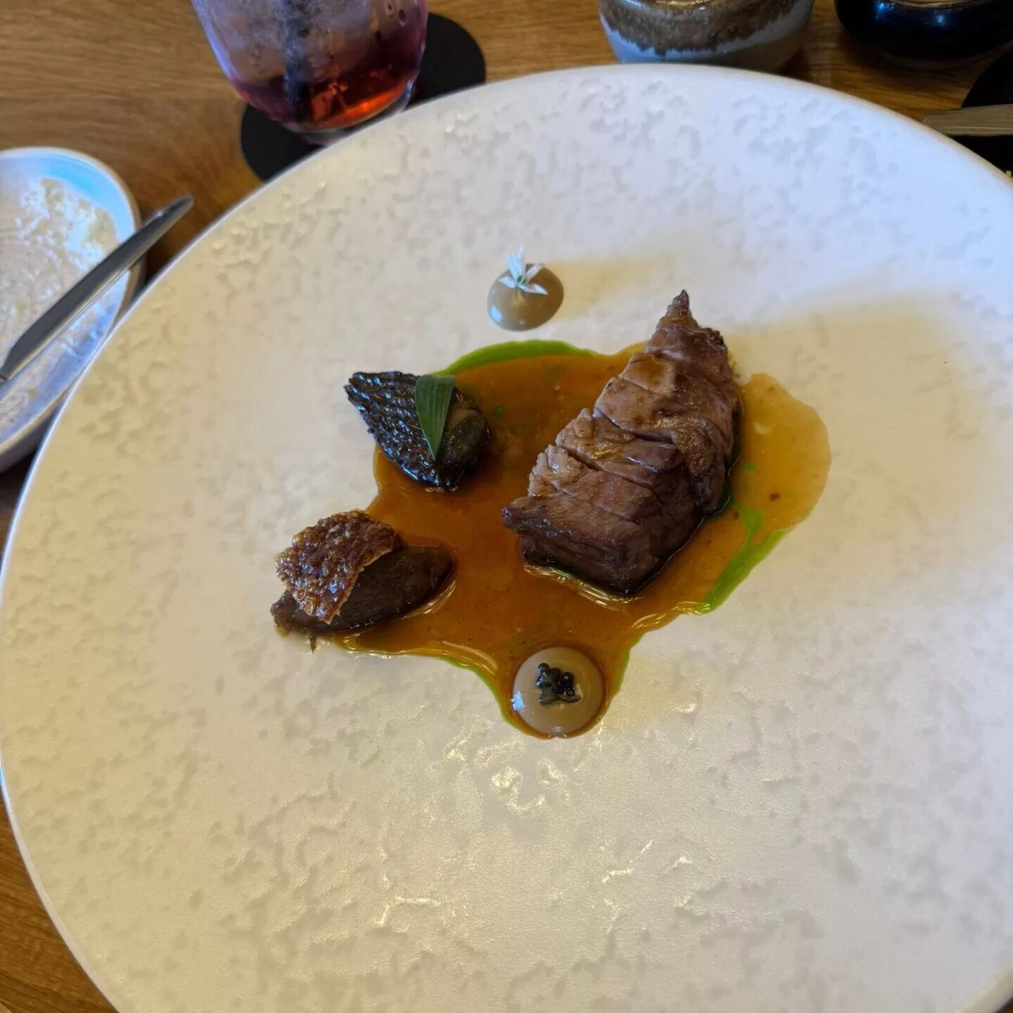 A plated dish featuring tender roast pork rib-eye slices, accompanied by small pieces of crispy pork, green sauce, and garnishes, served on a textured white plate.