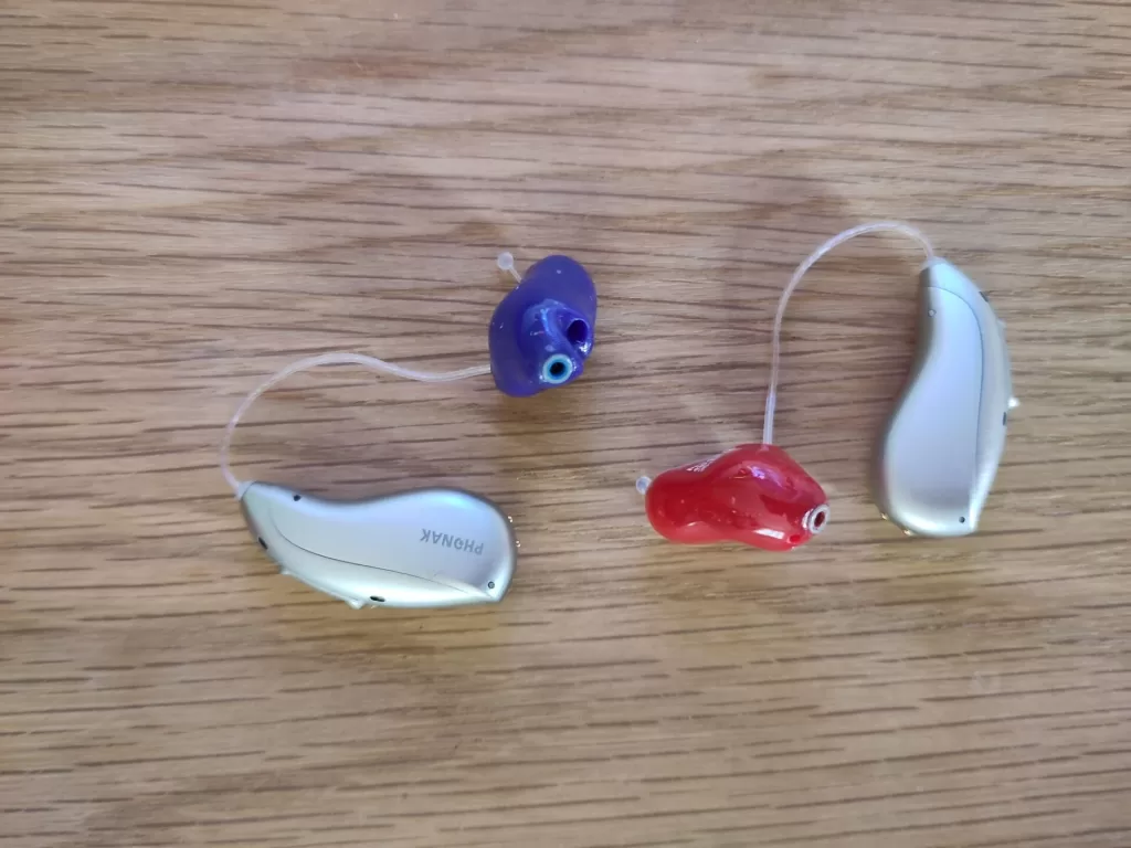 A close-up image of two Phonak Audéo Sphere Infinio I90 hearing aids, one with a purple ear mould and one with a red ear mould, alongside a silver device, all resting on a wooden surface.