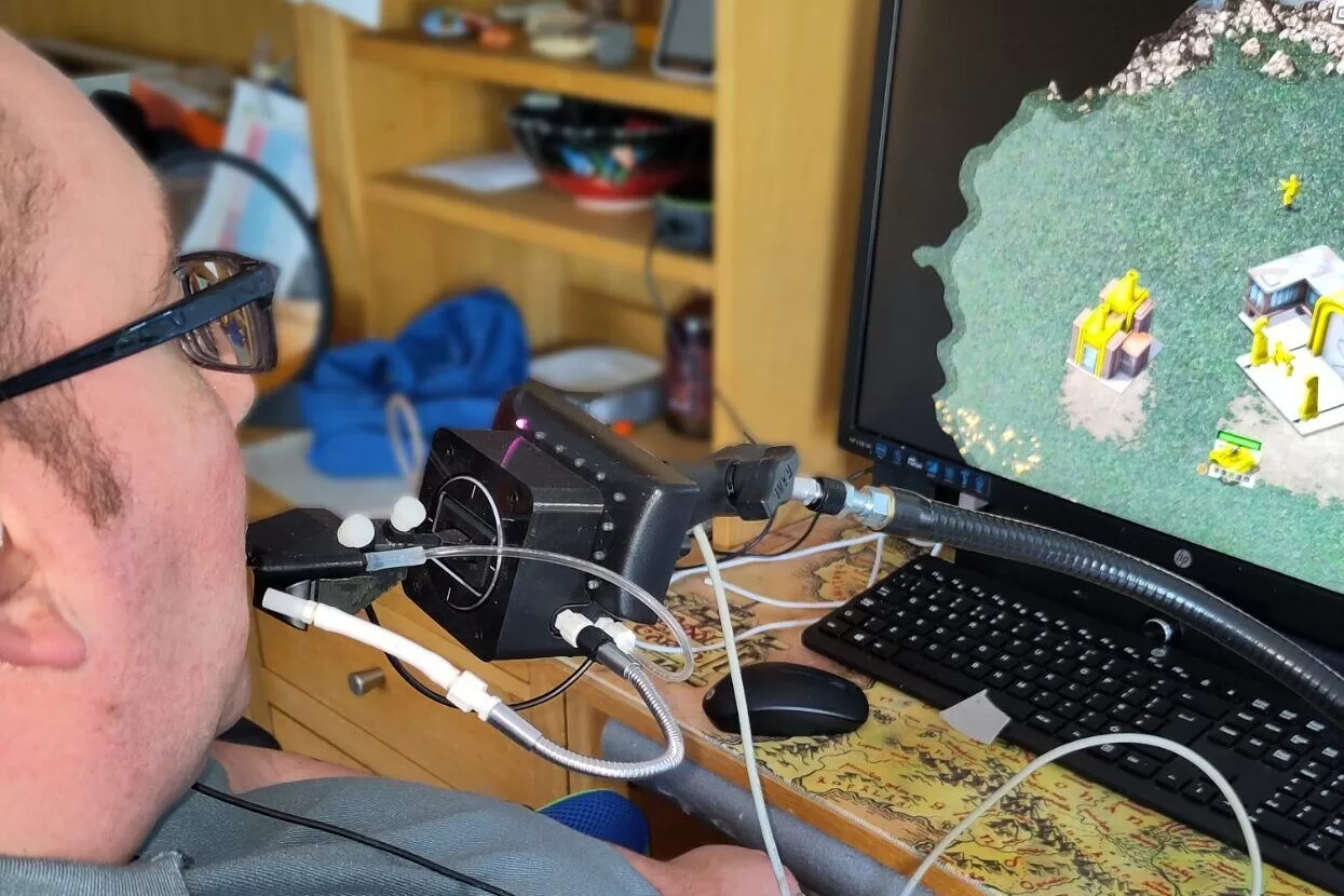 A person using a mouth-operated Quadstick adaptive game controller while playing a video game on a computer monitor.