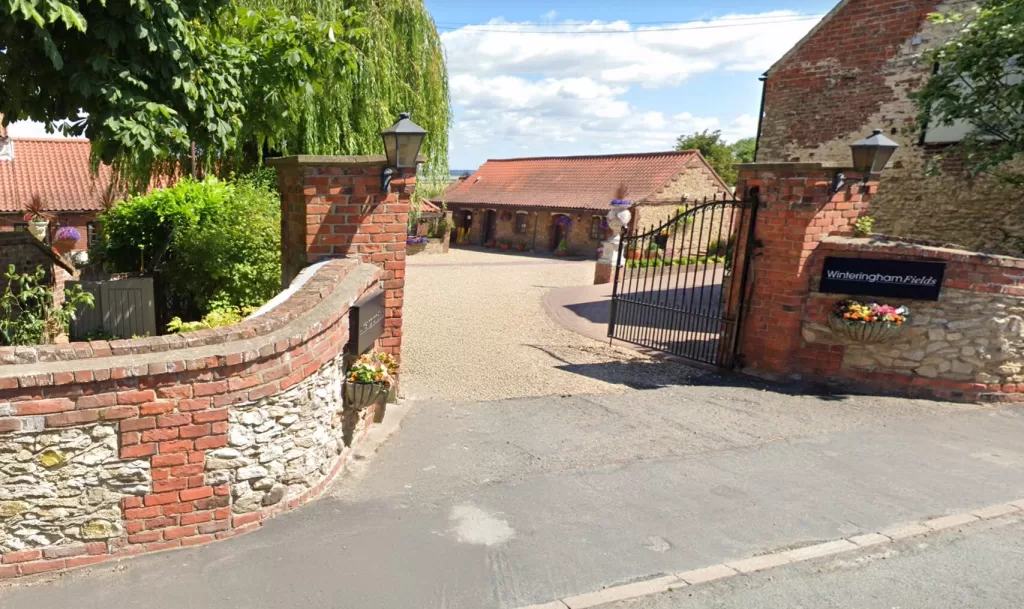 Entrance gate to Winteringham Fields restaurant, featuring a brick wall, a metal gate, and lush greenery, under a clear blue sky.