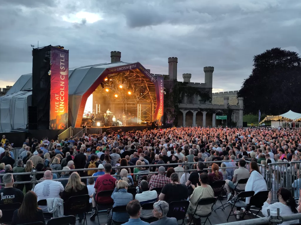 A large crowd gathers in front of a stage during a live concert at Lincoln Castle, with performers visible on stage and historic castle architecture in the background under a cloudy sky.