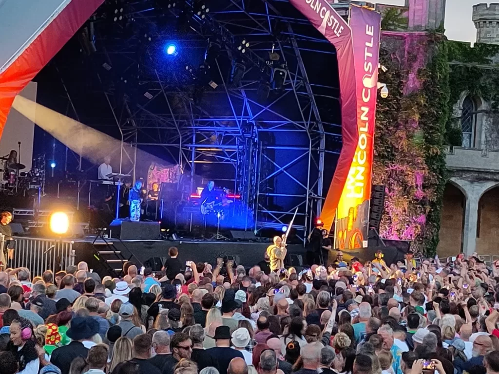 A vibrant outdoor concert scene at Lincoln Castle featuring the band James performing on stage in front of an enthusiastic crowd, with colourful lighting and decorations.