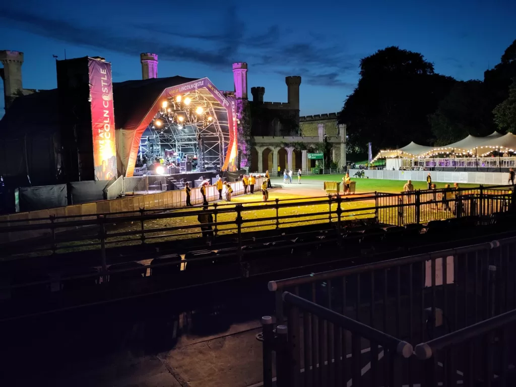 A view of the concert stage at Lincoln Castle, illuminated at night with colorful lights, surrounded by a grassy area and temporary structures.