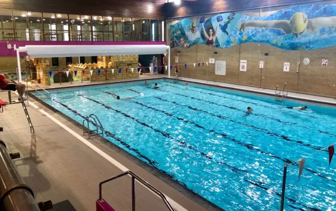 A wide view of the swimming pool at One NK Leisure Centre, featuring swimmers in lanes, colorful lane dividers, and a lifeguard on duty.