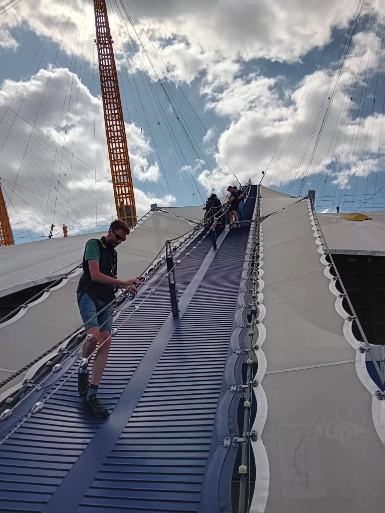 Climbers ascending the steep walkway on the O2 Arena under a partly cloudy sky.