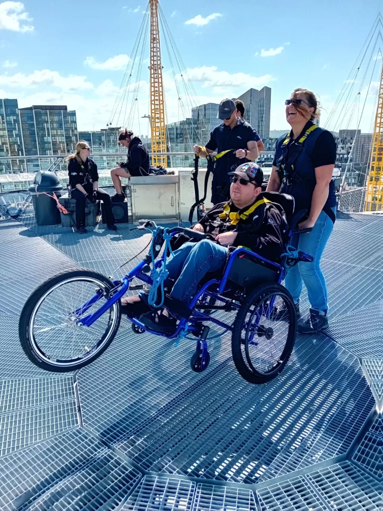 A person in a wheelchair being assisted by staff on the Platform at the top of the O2 Arena, with a bright blue sky and cityscape in the background.