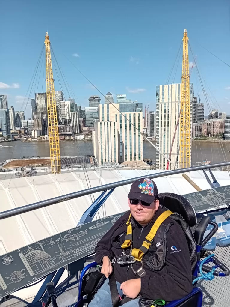 Person wearing a harness and sitting in a manual wheelchair on top of the O2 Arena, with a view of the London skyline in the background.