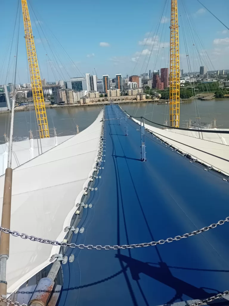 View from the top of the O2 Arena in London, showing the walkway and city skyline in the background under a bright blue sky.