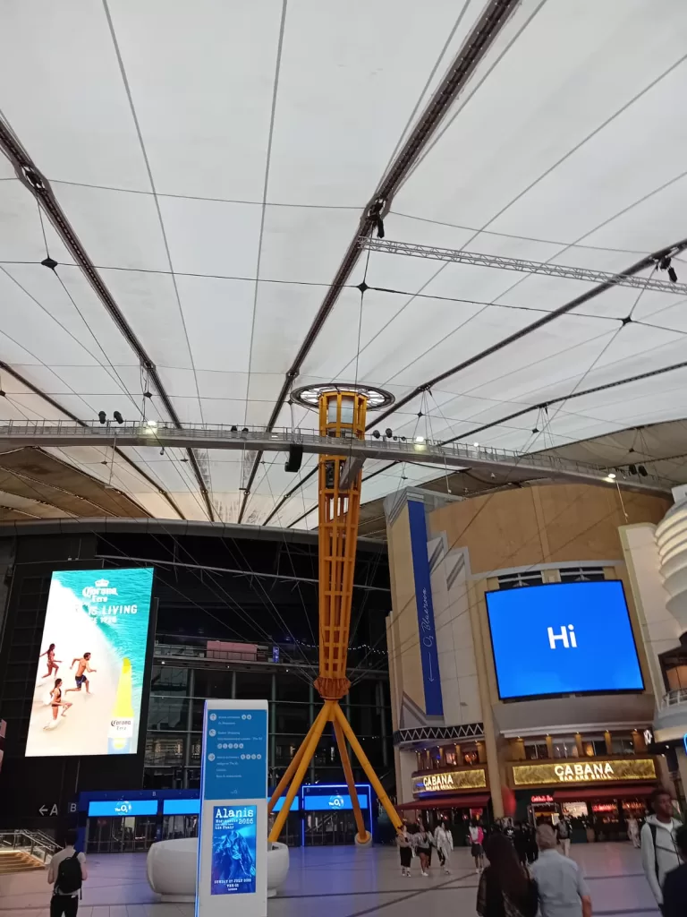 Interior view of the O2 Arena, featuring a high ceiling with a yellow support structure, large screen advertisements, and a spacious area filled with visitors.