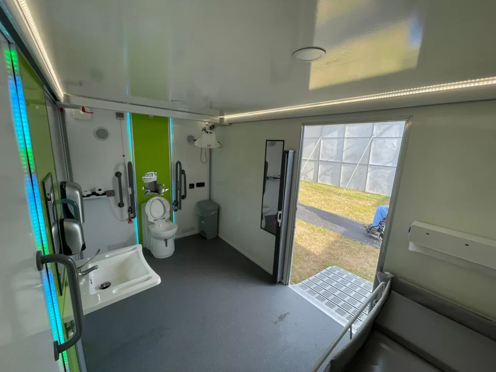 Interior of a Mobiloo with a toilet, sink, and grab rails, and a ceiling hoist designed for accessibility at outdoor events.