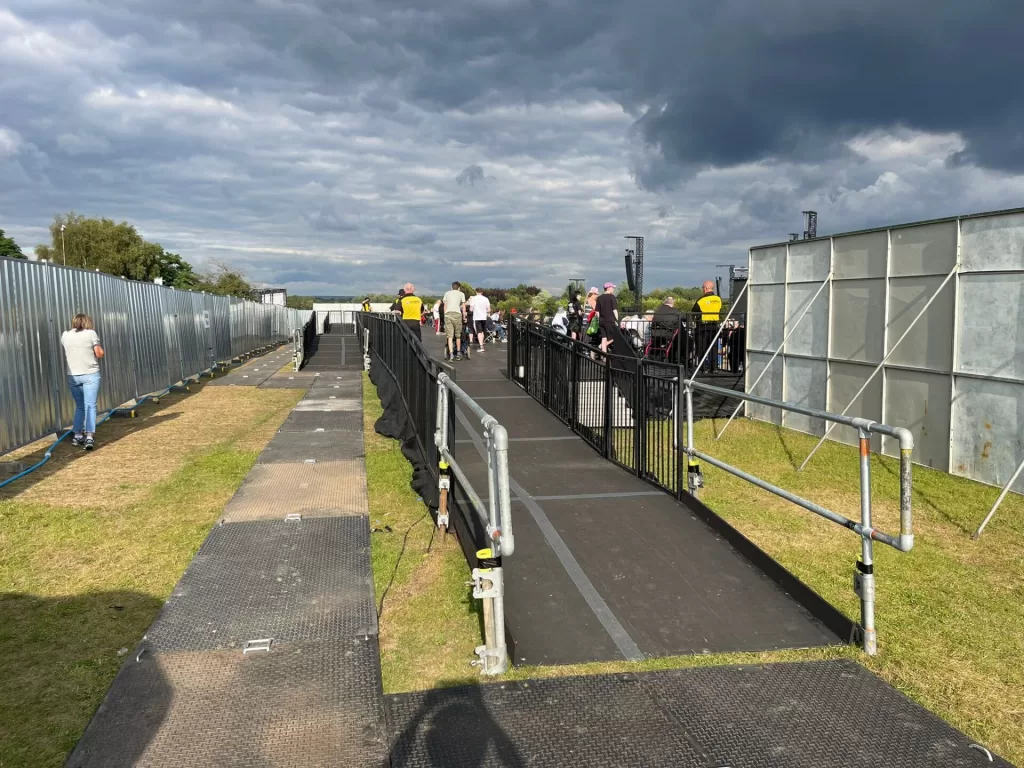 A smooth, accessible walkway leading to a viewing platform at Heaton Park during an outdoor concert, with people walking along the path.