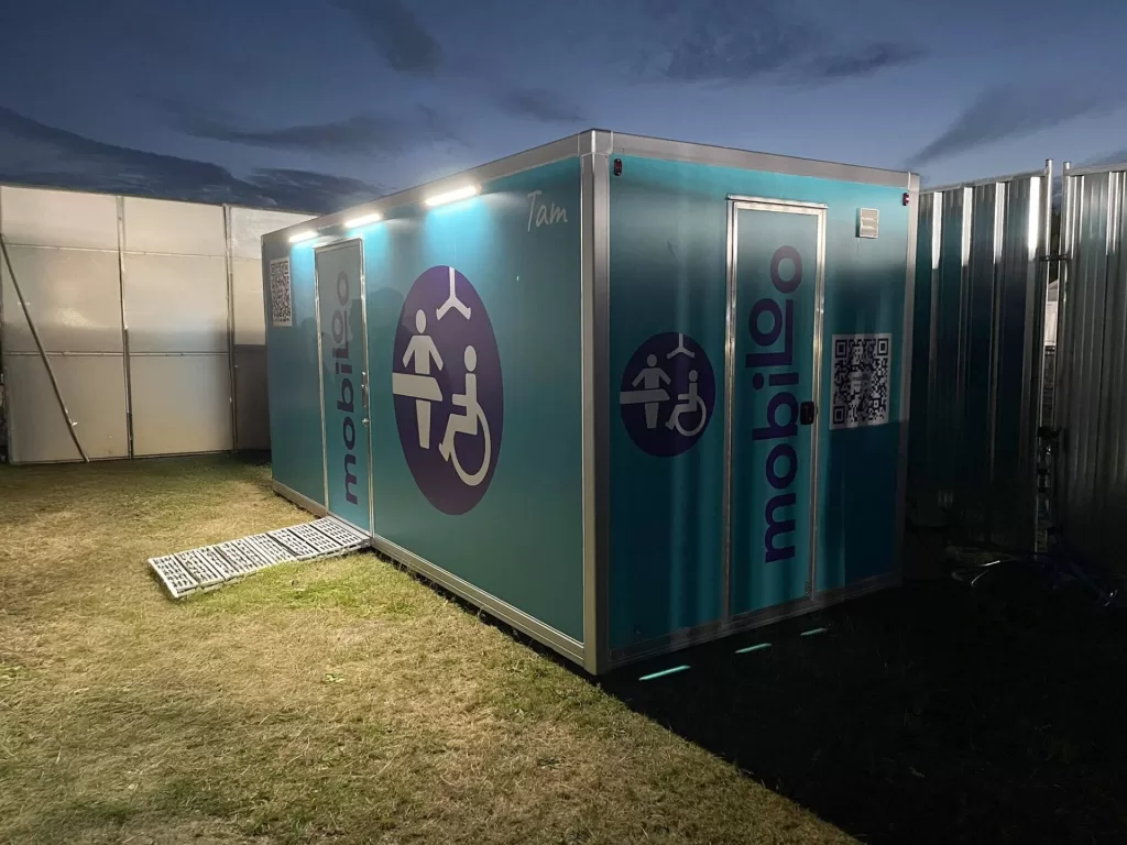 A Mobiloo accessible toilet facility at an outdoor event, illuminated at night. The building is blue with symbols indicating accessibility features, and there is a ramp leading to the entrance.