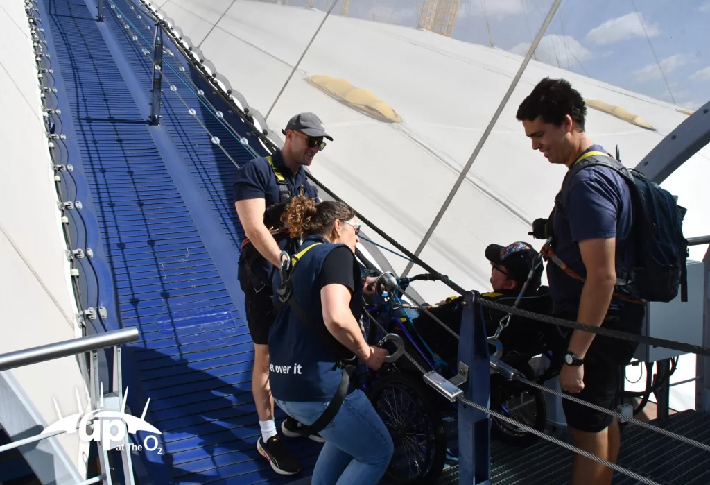 A group of four people preparing for a climb at the O2 Arena, with one person in a wheelchair being assisted by staff on a blue walkway.