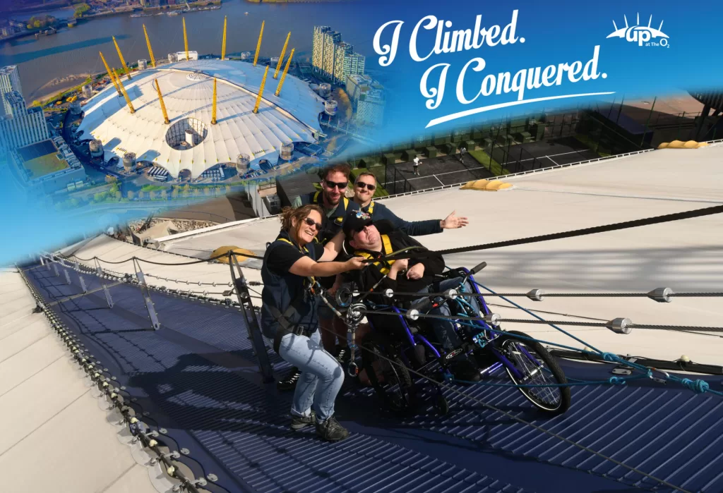 Group of four people on top of the O2 Arena, celebrating their climb with smiles. One person is in a manual wheelchair, secured with a harness, while the others pose enthusiastically. Background shows the arena's unique architecture and a blue sky.