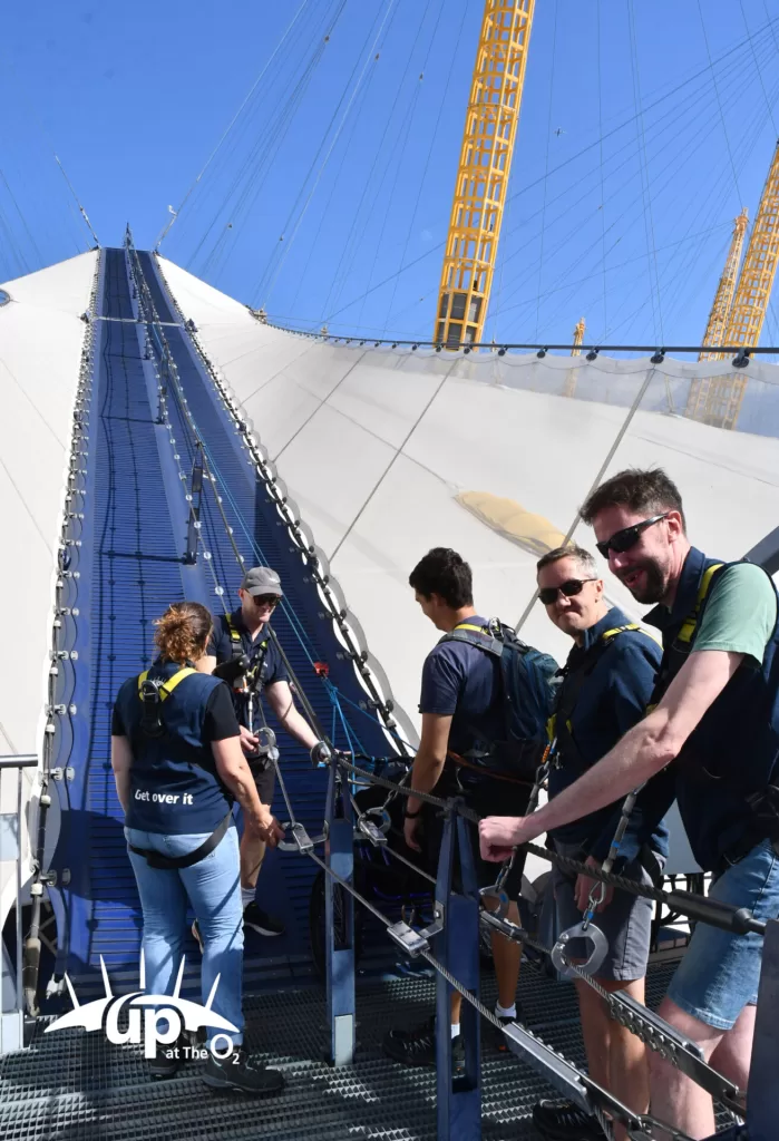 Group participating in the 'Up at the O2' climb, harnessed and preparing to ascend the O2 Arena under a clear blue sky.