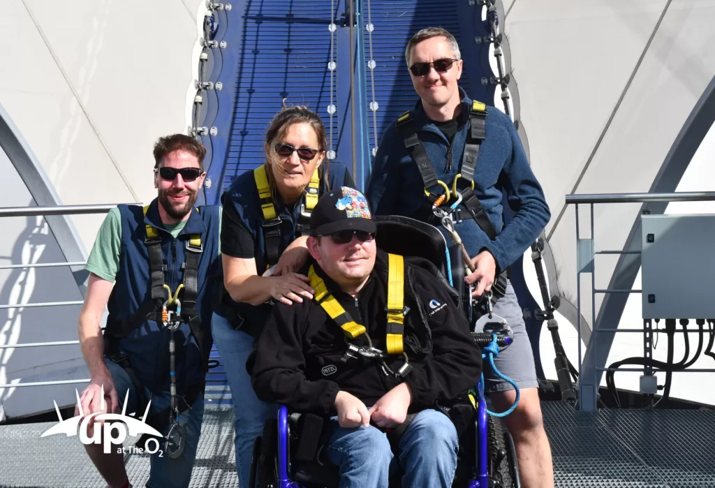 Group of four friends preparing for the climb at Up at The O2, with one person in a wheelchair wearing a harness and smiling.