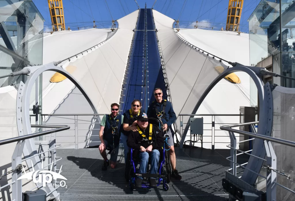 A group of four people preparing to climb at the O2 Arena, with one person in a wheelchair and harness, standing on a walkway with the arena's iconic dome structure in the background.