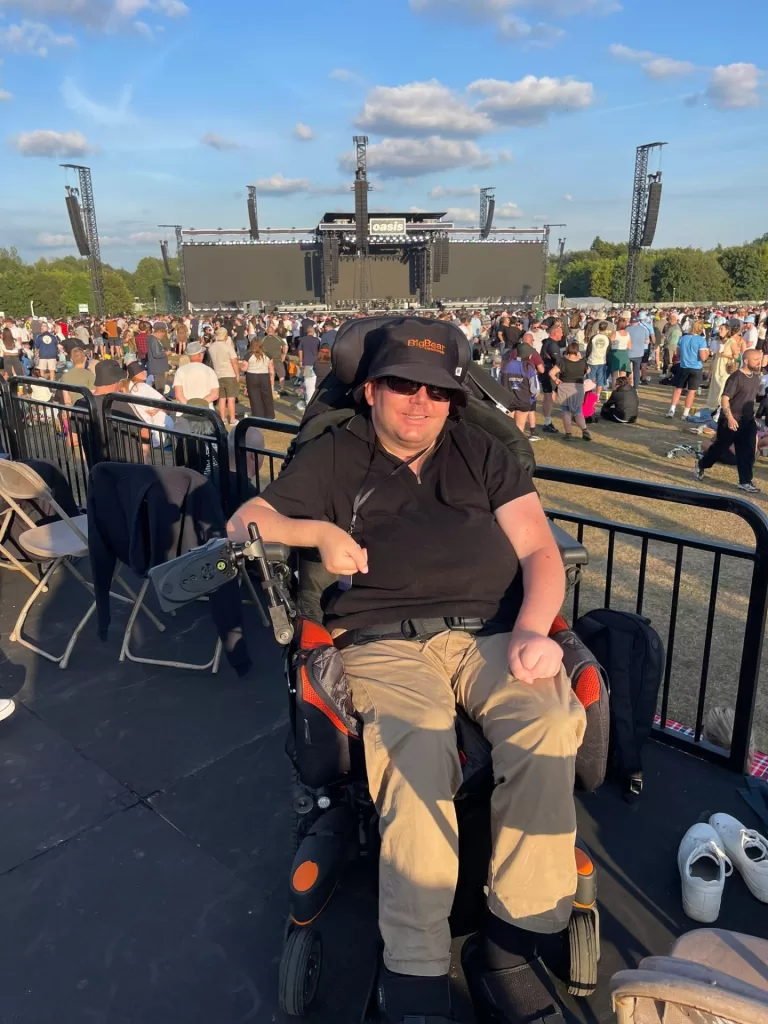 A person in a powered wheelchair is seated in front of a stage at an outdoor concert, with a large crowd in the background.