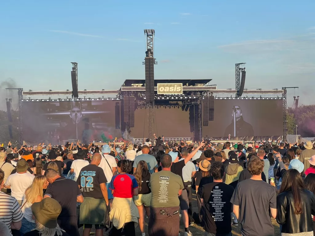 An outdoor concert scene at Heaton Park with a large crowd watching a performance on stage by Oasis, featuring a prominent video screen and a clear blue sky.