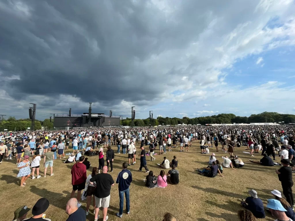 A large crowd gathered in Heaton Park for an Oasis gig, with a stage and sound equipment visible in the background under a cloudy sky.