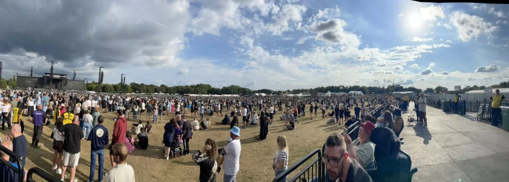 A panoramic view of a large crowd at an outdoor concert in Heaton Park, Manchester, with a stage in the distance and a clear sky.