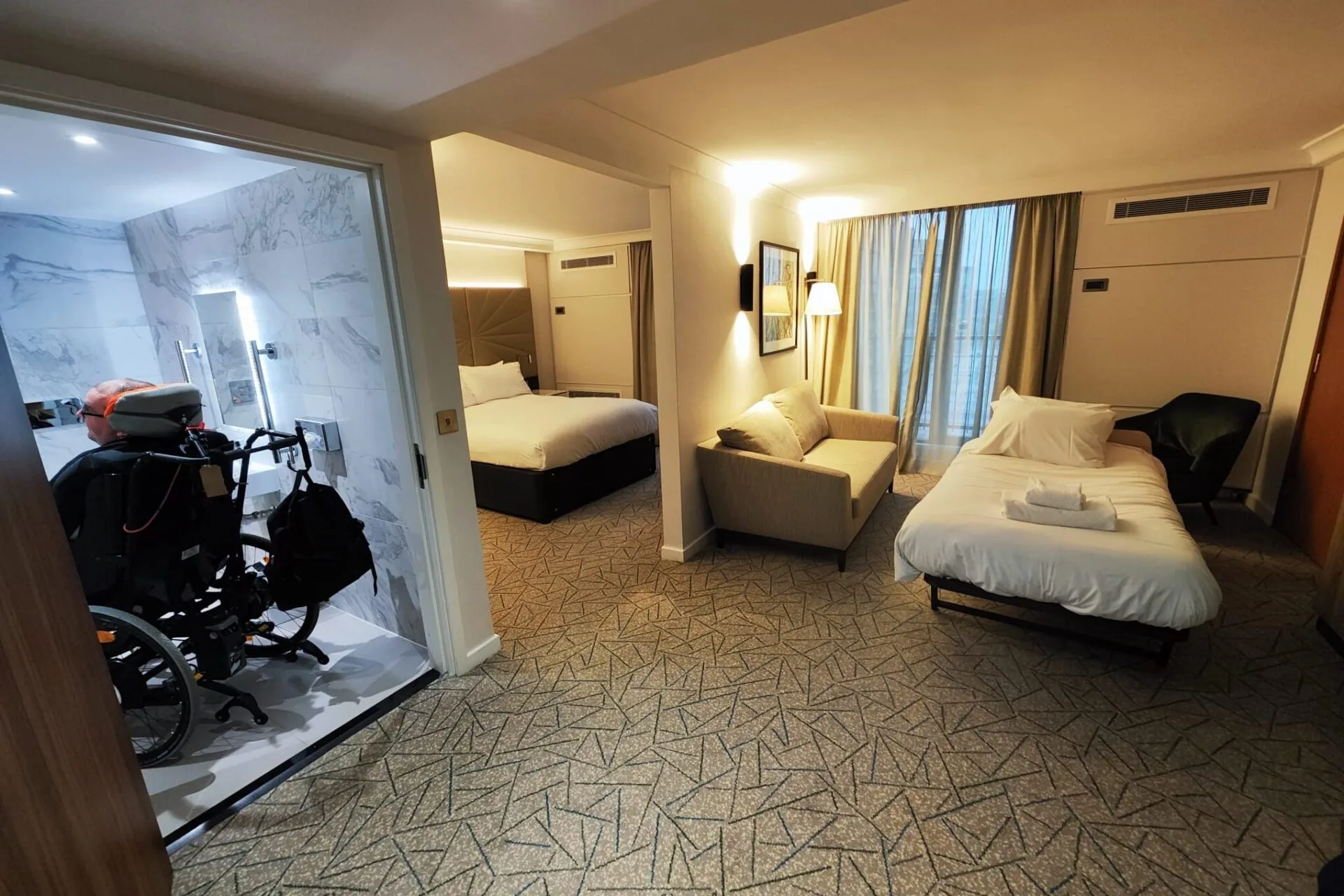 A spacious hotel room,  With a wheelchair in the bathroom on the left and a fold up bed on the right of the picture